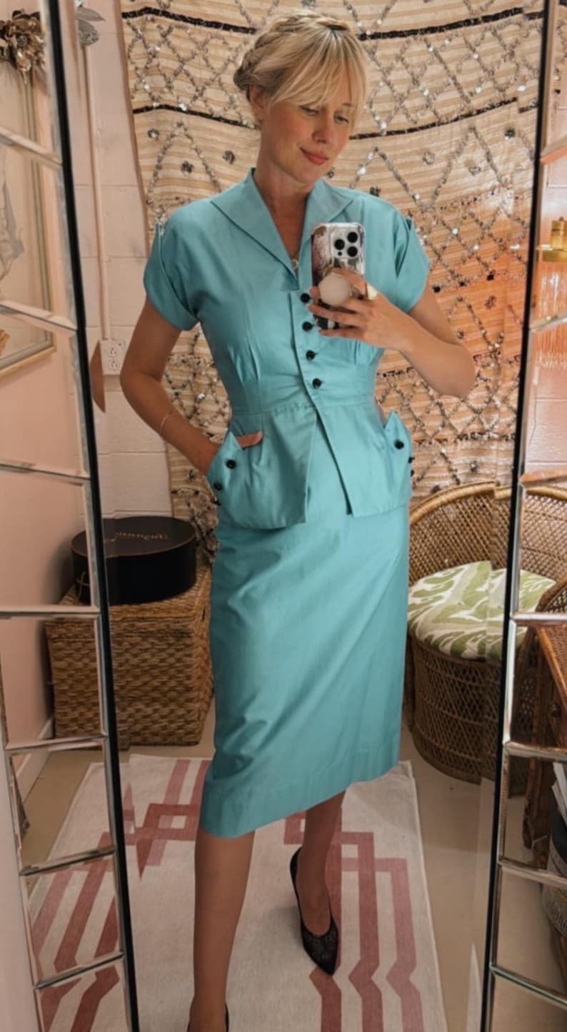 Pode incluir: Um conjunto de duas pe&ccedil;as azul claro em estilo vintage. Inclui um top com gola e mangas curtas com bot&otilde;es pretos e uma saia a condizer at&eacute; ao joelho. O conjunto &eacute; completado com sapatilhas pretas. A est&eacute;tica geral &eacute; retro e elegante.