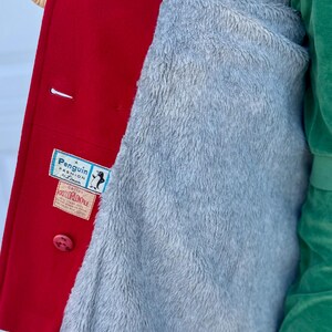 K&ouml;nnte beinhalten: Ein roter Wollmantel mit grauem, flauschigem Futter. Der Mantel hat einen Knopfverschluss und ein Etikett mit der Aufschrift "Penguin Fashion by Lyman". Das Futter ist ein weiches, pl&uuml;schiges Material.