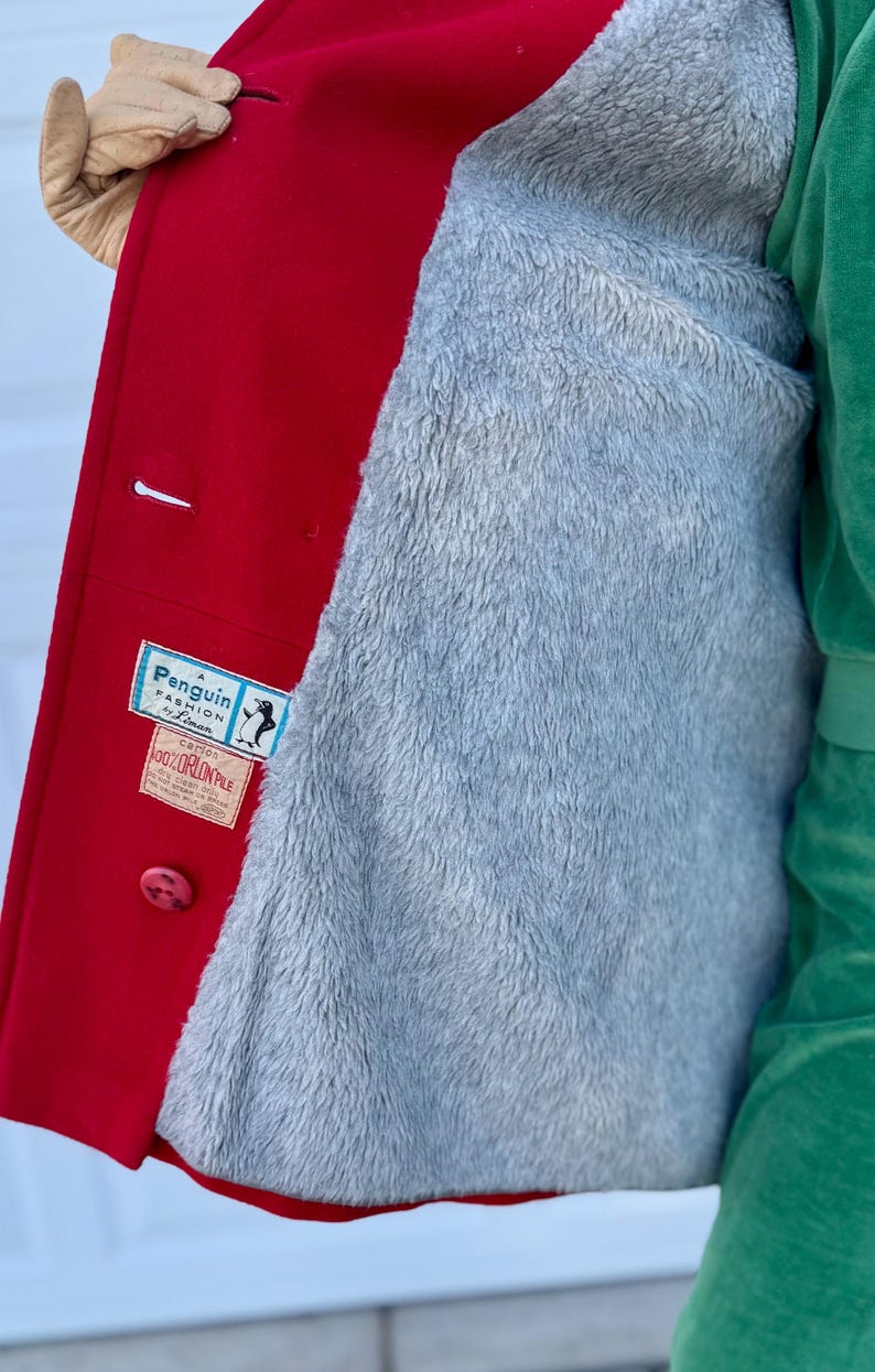K&ouml;nnte beinhalten: Ein roter Wollmantel mit grauem, flauschigem Futter. Der Mantel hat einen Knopfverschluss und ein Etikett mit der Aufschrift "Penguin Fashion by Lyman". Das Futter ist ein weiches, pl&uuml;schiges Material.