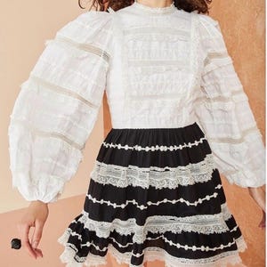 May include: A white blouse with long, ruffled sleeves and a high neckline. The blouse is paired with a black skirt featuring white lace trim and floral accents. The skirt has a ruffled hemline.