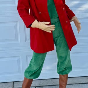 K&ouml;nnte beinhalten: Eine Person tr&auml;gt einen roten, zweireihigen Mantel, eine gr&uuml;ne Samthose und braune Lederstiefel. Das Outfit umfasst ein gr&uuml;nes Oberteil und beige Handschuhe. Ein Pelzhut vervollst&auml;ndigt den Look.