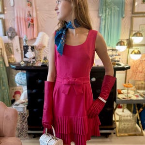 May include: A pink sleeveless dress with a bow at the waist and a pleated skirt. The dress is paired with white gloves and a white wicker handbag.