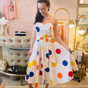 May include: A strapless white midi dress featuring a pattern of large, multicolored polka dots in red, blue, yellow, and orange. The dress includes a matching fabric tie-waist belt and a full, flared skirt that falls below the knee.