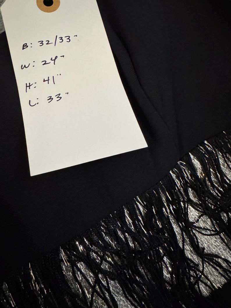 Pode incluir: Pe&ccedil;a de vestu&aacute;rio preta com franjas prateadas. Uma etiqueta mostra as medidas: B: 81/84 cm, L: 61 cm, A: 104 cm, C: 84 cm. As franjas adicionam textura e interesse visual &agrave; borda do tecido.