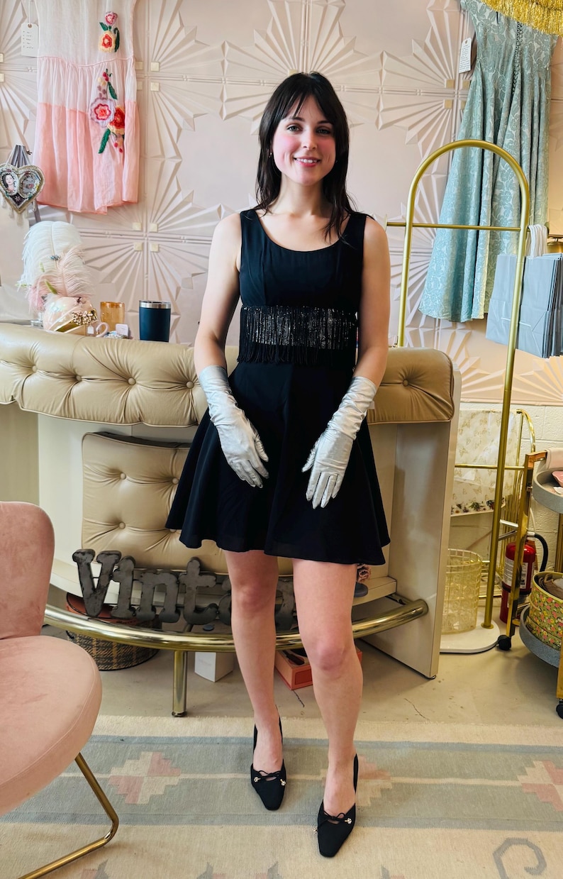Pode incluir: Um vestido preto sem mangas com cintura com franjas. O vestido &eacute; combinado com luvas prateadas e saltos pretos. O fundo apresenta uma loja de estilo vintage com elementos decorativos e roupas.