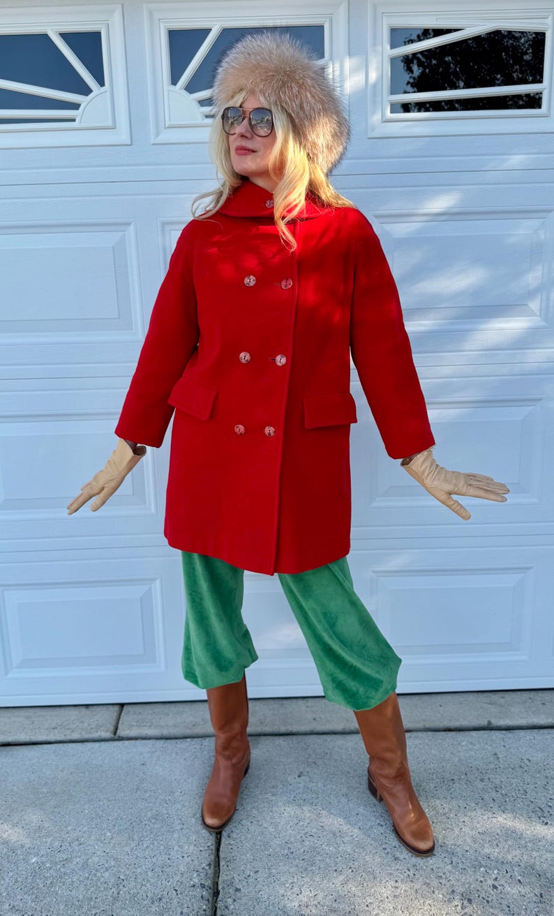 K&ouml;nnte beinhalten: Eine Person tr&auml;gt einen roten, zweireihigen Mantel, eine gr&uuml;ne Samthose, braune Lederstiefel und einen Pelzhut. Der Mantel hat gro&szlig;e Kn&ouml;pfe und ein klassisches Design. Das Outfit wird durch beige Handschuhe und eine Sonnenbrille erg&auml;nzt.