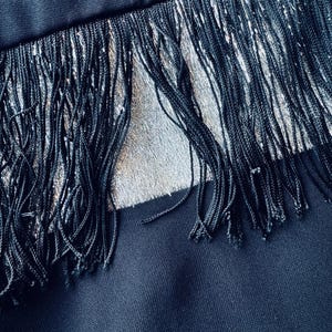 Pode incluir: Close-up de um tecido preto com detalhes de franjas prateadas. As franjas s&atilde;o feitas de fios longos e finos, criando um efeito texturizado. O tecido parece ser azul marinho escuro, com um brilho sutil.