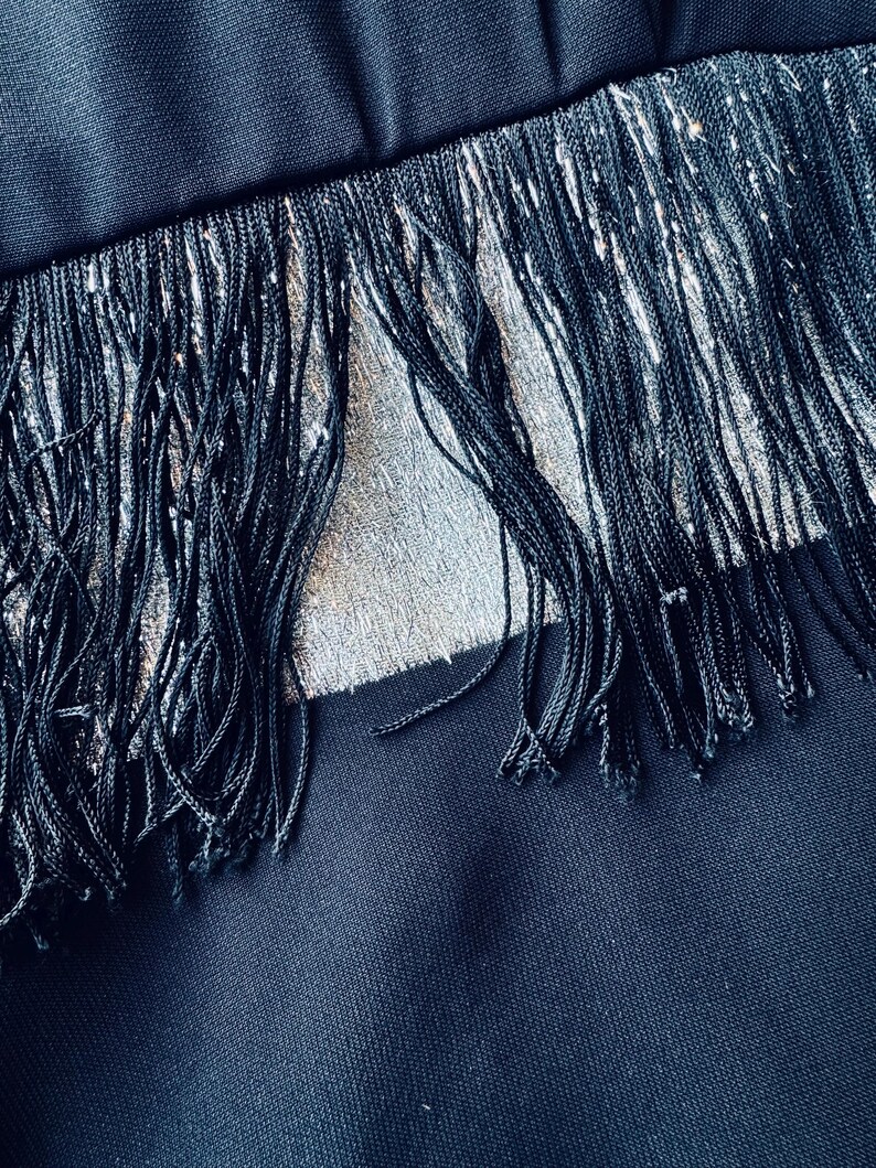 Pode incluir: Close-up de um tecido preto com detalhes de franjas prateadas. As franjas s&atilde;o feitas de fios longos e finos, criando um efeito texturizado. O tecido parece ser azul marinho escuro, com um brilho sutil.