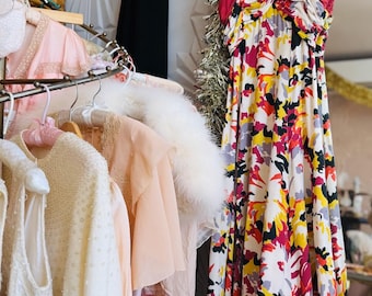May include: A rack of vintage clothing, including a colorful floral print dress with a red bodice. Other items include a white beaded dress, a pink jacket, and a black dress. A white fur stole is also visible.