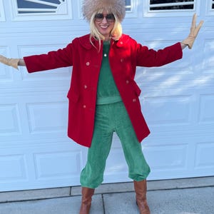K&ouml;nnte beinhalten: Eine Frau tr&auml;gt einen roten, zweireihigen Mantel, eine gr&uuml;ne Samthose und braune Stiefel. Sie tr&auml;gt auch einen Pelzhut und eine Sonnenbrille. Das Outfit wird durch beige Handschuhe erg&auml;nzt.