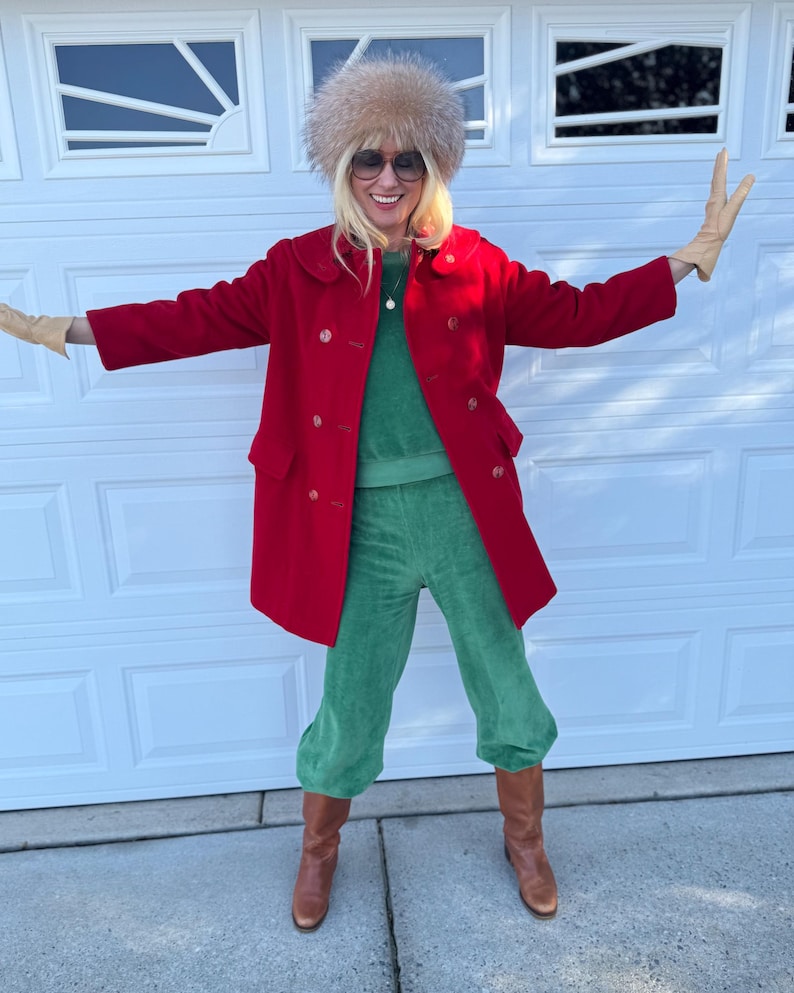 K&ouml;nnte beinhalten: Eine Frau tr&auml;gt einen roten, zweireihigen Mantel, eine gr&uuml;ne Samthose und braune Stiefel. Sie tr&auml;gt auch einen Pelzhut und eine Sonnenbrille. Das Outfit wird durch beige Handschuhe erg&auml;nzt.