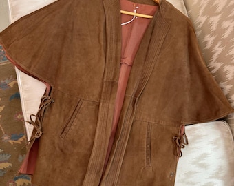 May include: A brown suede jacket with a cape-like design, hanging on a wooden hanger. The jacket features a notched collar, front pockets, and tie closures on the sides. The interior lining is a muted pink color.