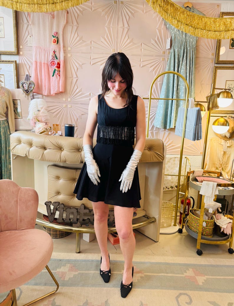 Pode incluir: Um vestido preto sem mangas com franjas na cintura. O vestido &eacute; combinado com luvas brancas e saltos pretos. O fundo inclui decora&ccedil;&atilde;o vintage, uma cadeira rosa e franjas douradas.