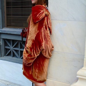 Könnte beinhalten: Ein rostfarbenes Samtkleid mit einer großen Schleife auf der Schulter. Das Kleid hat lange Ärmel und einen drapierten Rock. Die Person trägt einen weißen Hut mit braunem Akzent und passende Absätze. Das Outfit ist elegant und von Vintage inspiriert.