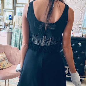 Pode incluir: Vestido preto com detalhe de la&ccedil;o nas costas e acabamento com franjas. O vestido &eacute; sem mangas e tem uma saia rodada. Um len&ccedil;o preto com detalhes dourados &eacute; usado na cabe&ccedil;a.