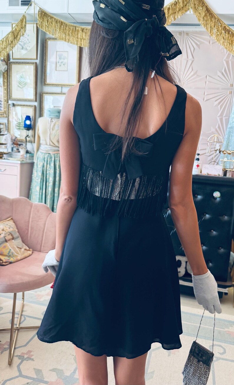 Pode incluir: Vestido preto com detalhe de la&ccedil;o nas costas e acabamento com franjas. O vestido &eacute; sem mangas e tem uma saia rodada. Um len&ccedil;o preto com detalhes dourados &eacute; usado na cabe&ccedil;a.
