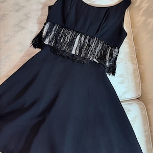 Pode incluir: Um vestido preto sem mangas com um corpete justo e uma saia rodada. O corpete apresenta franjas na regi&atilde;o da cintura. O vestido &eacute; feito de um tecido fluido, e o estilo geral &eacute; elegante e adequado para ocasi&otilde;es formais.