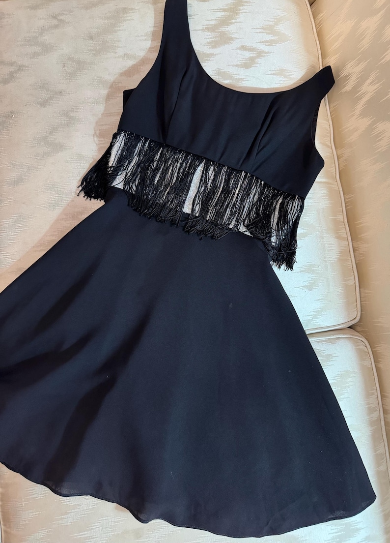 Pode incluir: Um vestido preto sem mangas com um corpete justo e uma saia rodada. O corpete apresenta franjas na regi&atilde;o da cintura. O vestido &eacute; feito de um tecido fluido, e o estilo geral &eacute; elegante e adequado para ocasi&otilde;es formais.