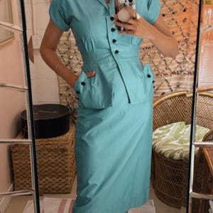 Pode incluir: Um conjunto de duas pe&ccedil;as azul claro em estilo vintage. A parte de cima tem mangas curtas, gola e bot&otilde;es pretos. A saia vai at&eacute; ao joelho. O conjunto &eacute; usado com sapatilhas pretas. A imagem &eacute; tirada num espelho.
