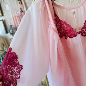 May include: A light pink vintage nightgown with sheer sleeves and a burgundy lace trim. The nightgown is hanging on a gold hanger. A white dress with floral embroidery is visible in the background.