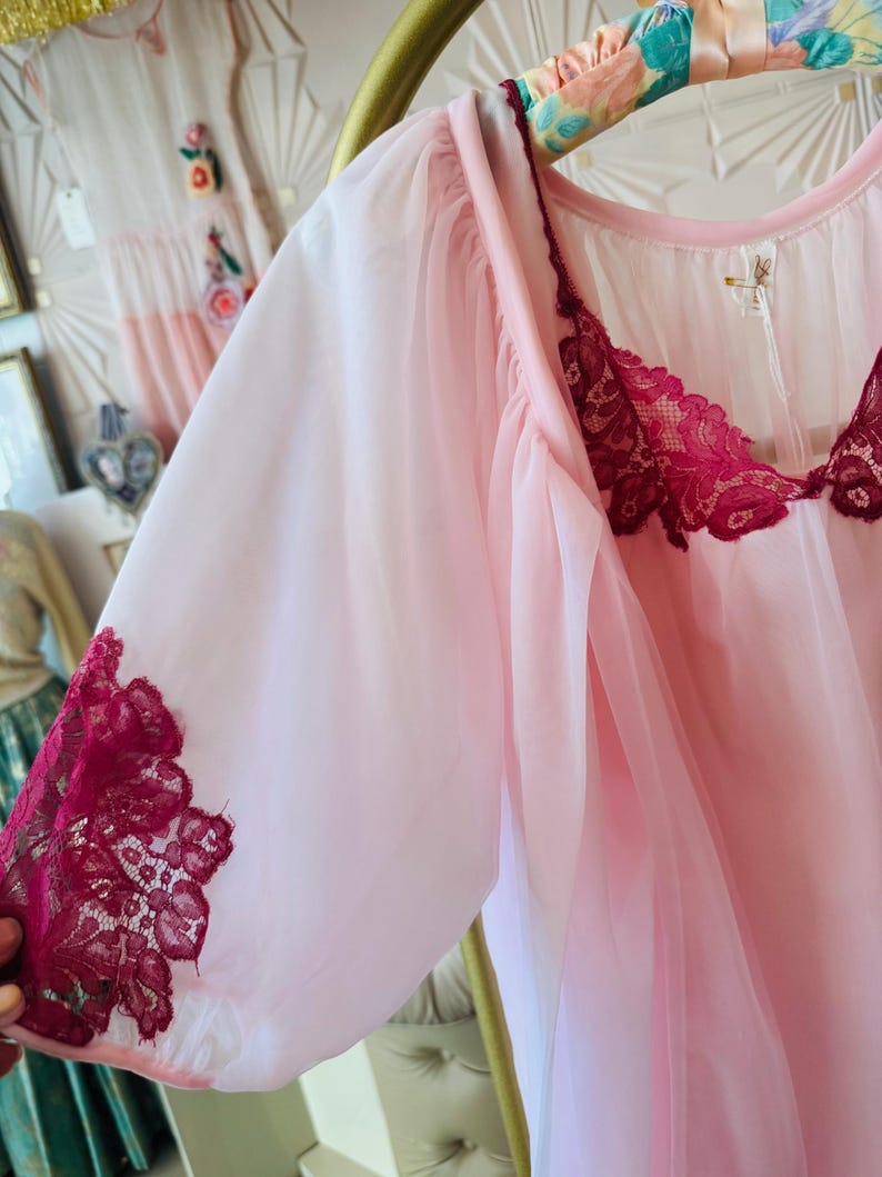 May include: A light pink vintage nightgown with sheer sleeves and a burgundy lace trim. The nightgown is hanging on a gold hanger. A white dress with floral embroidery is visible in the background.