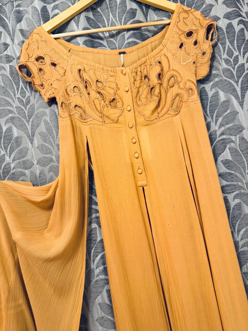 Może przedstawiać: Jasnopomarańczowa sukienka z kr&oacute;tkimi rękawami i zapięciem na guziki z przodu. Sukienka posiada skomplikowane kwiatowe hafty wok&oacute;ł dekoltu i rękaw&oacute;w. Sukienka wisi na drewnianym wieszaku.