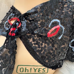 May include: A black sequined bolero jacket featuring a sheer mesh base and decorative floral-like accents in red and light blue. The garment is displayed on a mannequin with a green rectangular label at the base containing the text Oh!YES.