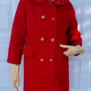 K&ouml;nnte beinhalten: Ein leuchtend roter Vintage-Mantel mit doppelreihigem Design. Der Mantel hat einen gro&szlig;en Kragen, zwei Vordertaschen und passende Kn&ouml;pfe. Die Person tr&auml;gt beige Handschuhe und eine gr&uuml;ne Hose.