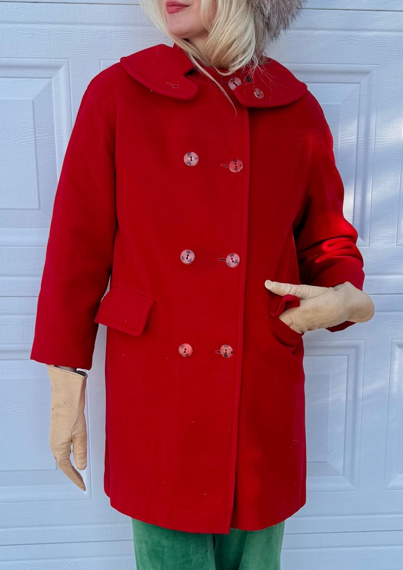 K&ouml;nnte beinhalten: Ein leuchtend roter Vintage-Mantel mit doppelreihigem Design. Der Mantel hat einen gro&szlig;en Kragen, zwei Vordertaschen und passende Kn&ouml;pfe. Die Person tr&auml;gt beige Handschuhe und eine gr&uuml;ne Hose.
