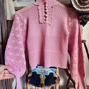 Puede incluir: Un suéter de ganchillo de manga larga en color rosa claro con cuello alto y una fila vertical de cinco botones a juego cubiertos de tela. La prenda presenta intrincados patrones florales en las mangas y un cuerpo de punto texturizado.