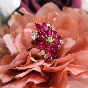May include: A gold ring featuring a cluster of marquise-cut deep pink stones arranged in a floral pattern, accented by small round clear stones set in gold prongs. The ring has a polished gold band and a multi-layered, dimensional design.