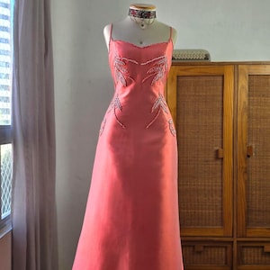 Puede incluir: Vestido de noche color coral con tirantes finos y falda larga. El corpiño presenta diseños bordados en forma de hojas. Se lleva un collar gargantilla a juego. El vestido se exhibe en un maniquí.
