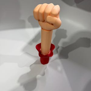 Op de afbeelding: Een licht perzikkleurig plastic object in de vorm van een gebalde vuist aan de bovenkant van een cilindrische steel. De steel is geplaatst in een felrode, geschulpte, bloemvormige voet die naar boven toe uitloopt en naar onderen toe versmalt.