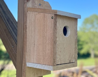 Puede incluir: Un nido de pájaros de madera con un agujero de entrada redondo, montado en un poste de madera. El nido está hecho de madera de color claro con un acabado natural. Incluye un pequeño voladizo de techo.