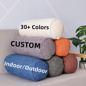 May include: A selection of cylindrical cushions in diverse hues, such as white, grey, orange, blue, and brown. The cushions display the text "30+ Colors", "CUSTOM", and "Indoor/Outdoor". They are arranged on a grey sofa.