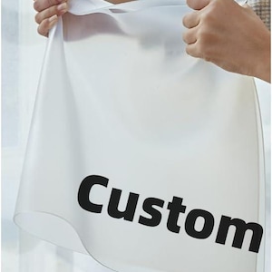 May include: A translucent, white sheet with the word "Custom" printed in bold, black letters. The sheet is being held up by two hands, showcasing its flexible and smooth texture. The background is a soft, neutral color.