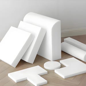 May include: An assortment of white foam shapes, including rectangular, cylindrical, and curved pieces. The collection features a variety of sizes and forms, suitable for various applications. The shapes are arranged on a light-colored wooden floor.