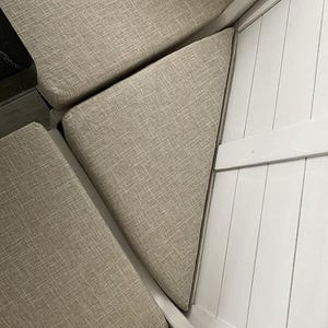 May include: Close-up of beige upholstered cushions arranged in a corner. The cushions have a textured fabric and are set against a white, paneled wooden structure. The arrangement suggests a built-in seating area.