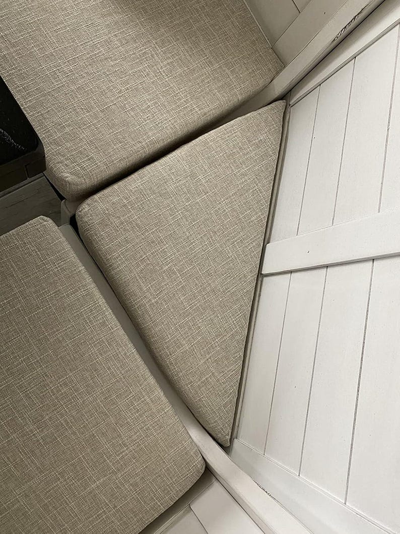 May include: Close-up of beige upholstered cushions arranged in a corner. The cushions have a textured fabric and are set against a white, paneled wooden structure. The arrangement suggests a built-in seating area.