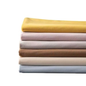 May include: A stack of folded fabric in various colors, including yellow, pink, gray, brown, beige, and blue. The fabrics appear to be soft and smooth, possibly blankets or sheets, arranged in a neat pile.