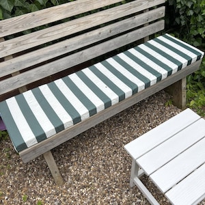 May include: A wooden garden bench with a green and white striped cushion. The bench is weathered and sits on a bed of small pebbles. A small white side table is next to the bench, with foliage in the background.