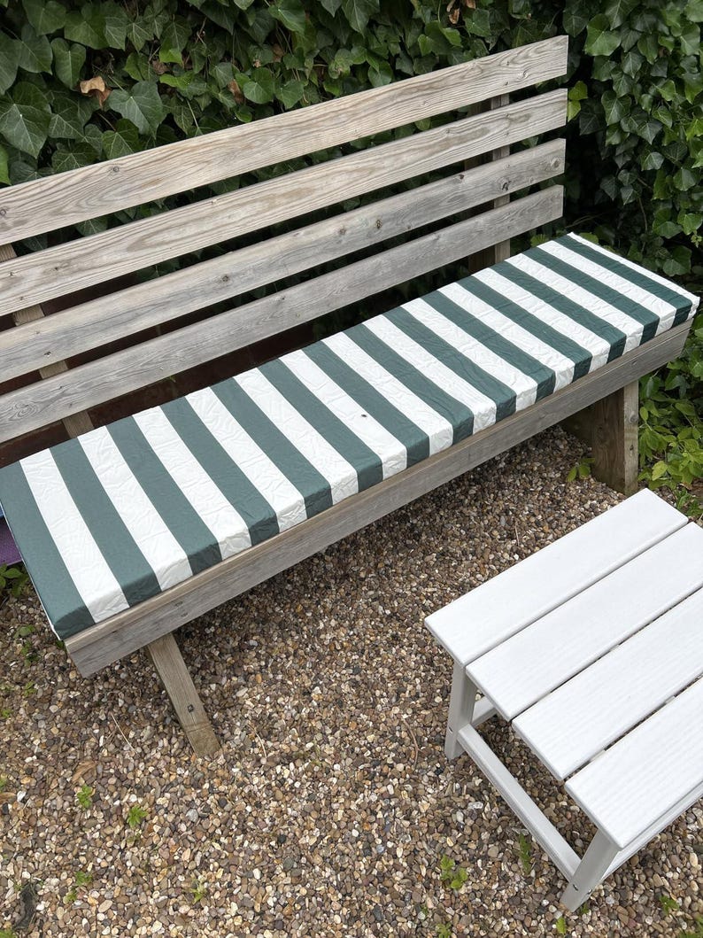 May include: A wooden garden bench with a green and white striped cushion. The bench is weathered and sits on a bed of small pebbles. A small white side table is next to the bench, with foliage in the background.