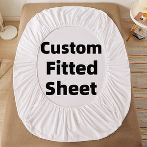 May include: A white fitted sheet with the words "Custom Fitted Sheet" in large black letters. The sheet is draped over a bed, showcasing its shape and design. The overall theme is bedding and home textiles.