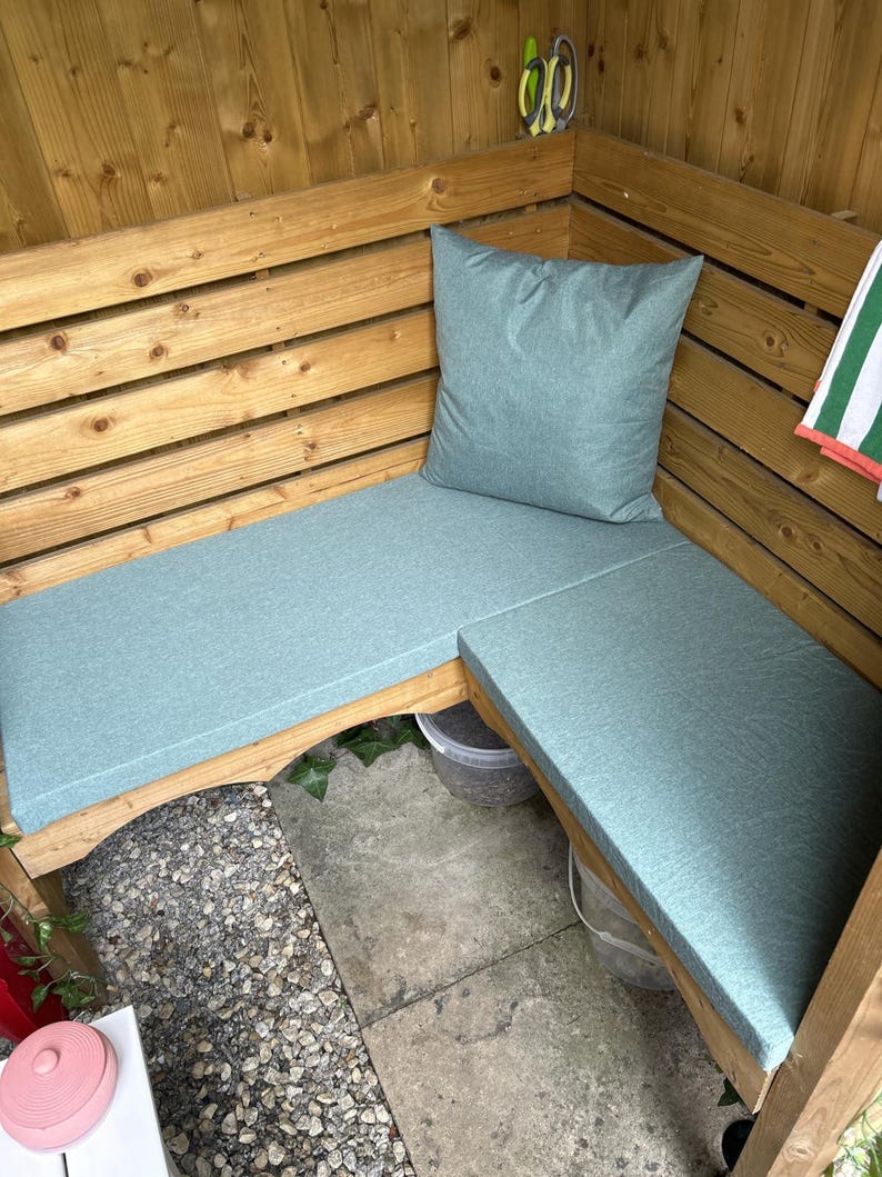 May include: A wooden outdoor bench with light blue cushions and a matching pillow. The bench is built into a wooden structure, creating a cozy seating area. A green and white striped towel hangs nearby.