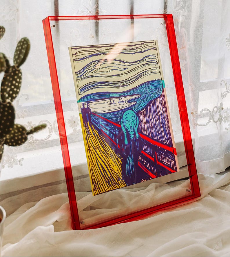 May include: A framed print of Edvard Munch's "The Scream" in a clear acrylic frame with red edges. The artwork features a figure with a wide open mouth against a backdrop of a bridge and a turbulent sky.