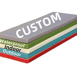 May include: A stack of custom-made, rectangular cushions with a gray top layer. The layers below are green, off-white, blue, and red. Text on the layers reads "Waterproof", "Indoor", and "Outdoor". The top layer has the word "CUSTOM" in white.
