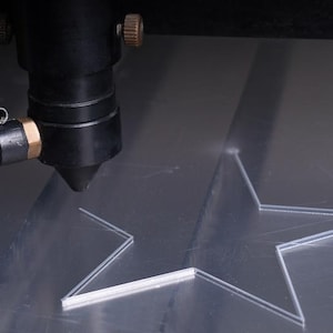 May include: A laser cutter precisely cutting a star shape into a clear acrylic sheet. The laser head is black with brass and blue fittings. The star is partially cut, showing the detail of the process.