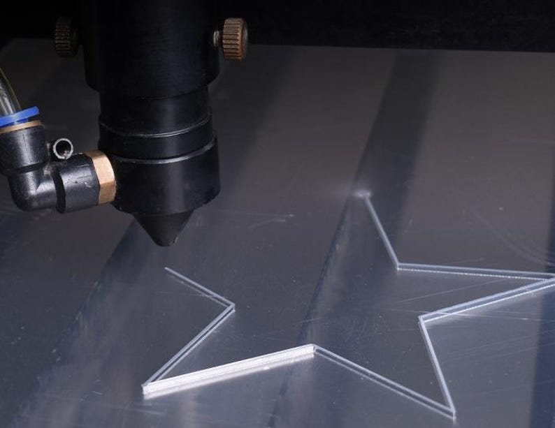 May include: A laser cutter precisely cutting a star shape into a clear acrylic sheet. The laser head is black with brass and blue fittings. The star is partially cut, showing the detail of the process.