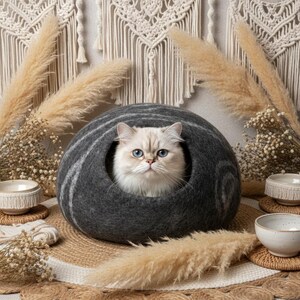 May include: A gray cat bed with a white and gray striped design, housing a fluffy white cat with blue eyes. The cat bed is surrounded by decorative elements, including dried plants and small ceramic bowls, creating a cozy and stylish pet accessory.