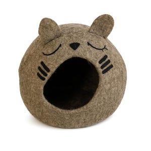 May include: A cat-shaped pet bed in a neutral brown color. The bed is made of felt and features a round shape with a large opening. Black details include closed eyes, a nose, and whiskers.
